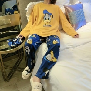 Mignon Pyjama Disney Donald pour femme jaune moutard et bleue porté par une femme qui s'assoit sur un lit dans une maison
