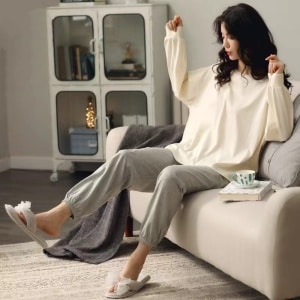 Pyjama d'été avec pull-over blanc manches chauve-souris et pantalon gris portée par une femme assise sur le canapé dans une maison