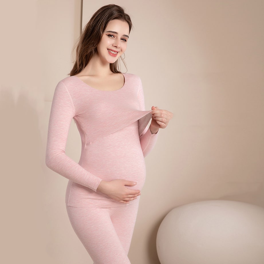 Femme brune enceinte qui porte un pyjama de grossesse rose, elle touche son ventre rond d'une main et de l'autre tire sur le haut de son pyjama