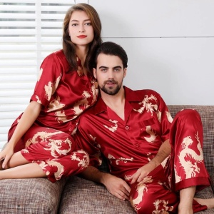 Un homme assis sur un canapé marron et une femme assise sur l'accoudoir derrière lui. Ils sont habillé en pyjama dragon sur fond rouge satiné